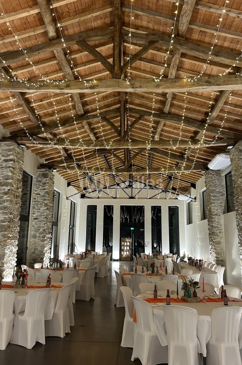 Salle de réception avec des tables décorées, des chaises blanches et un plafond en bois orné de guirlandes lumineuses.