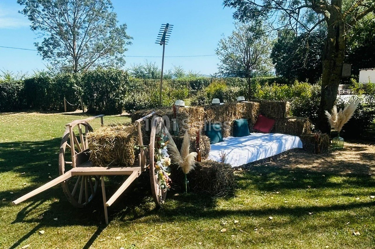 Banc rustique en bottes de paille avec coussins colorés, décor champêtre sous un arbre, près d'une charrette en bois.