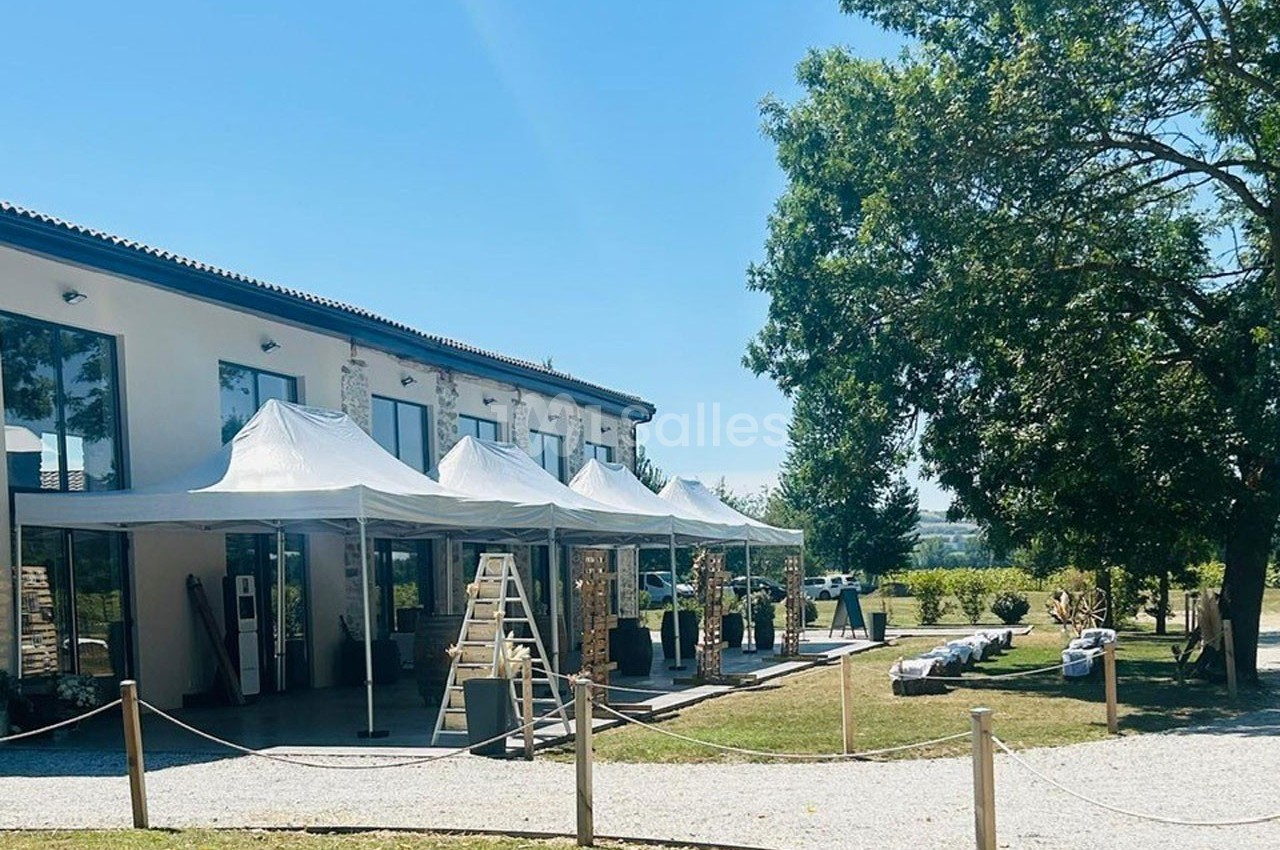 Extérieur d'un bâtiment moderne avec des tentes blanches, un chemin gravillonné et des arbres sous un ciel dégagé.