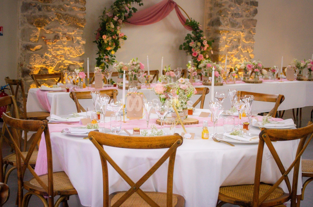 Salle décorée pour un mariage avec tables rondes, nappes blanches, chemins de table roses et arrangements floraux.