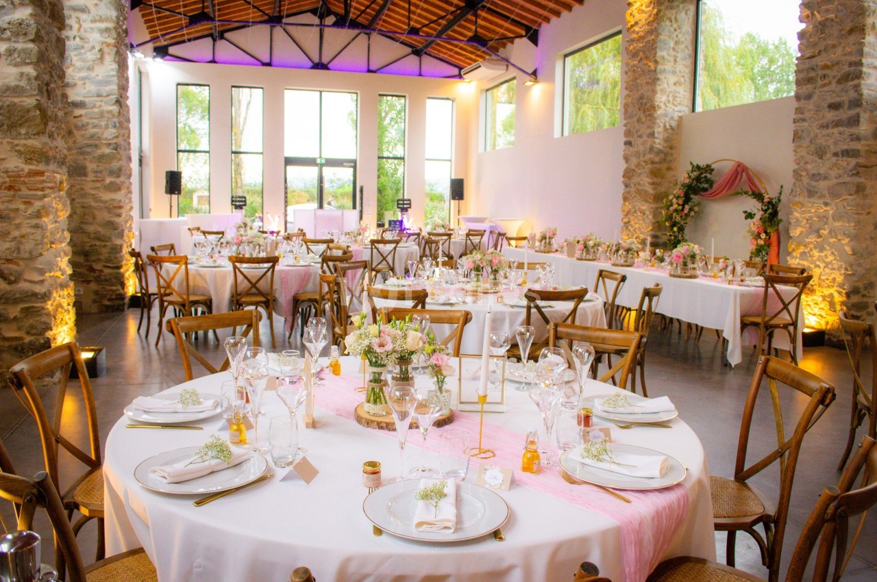 Salle de réception lumineuse avec tables décorées de fleurs et nappes blanches, entourées de chaises en bois.