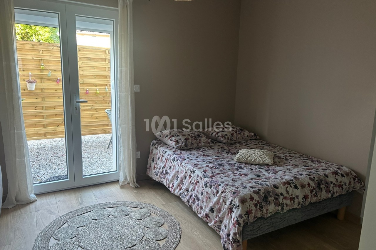 Chambre avec un lit double à motif floral, tapis rond au sol et porte-fenêtre donnant sur une terrasse en bois.