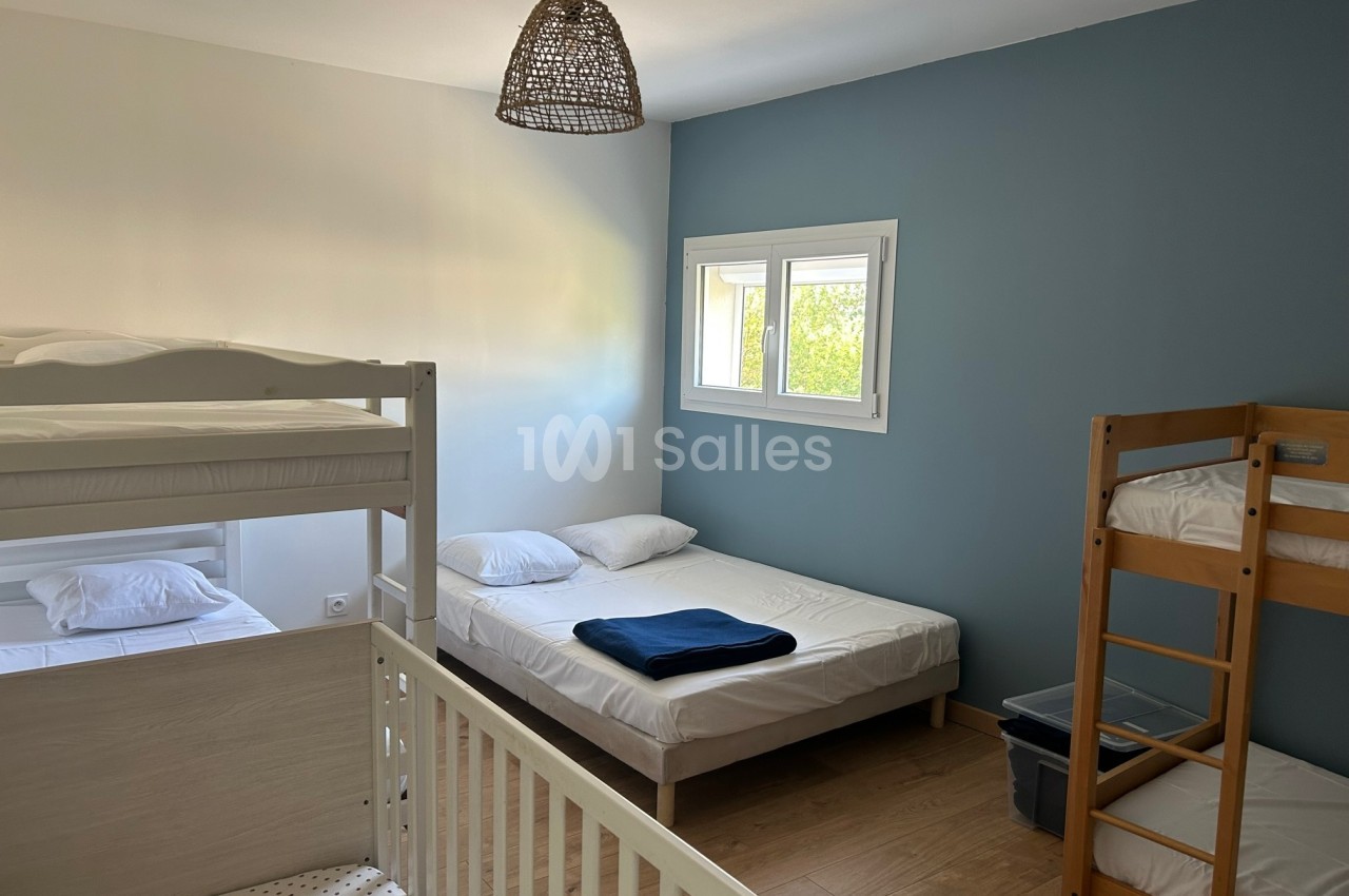 Chambre avec lits superposés, lits simples, lit bébé, murs blancs et bleus, et lumière naturelle par une fenêtre.