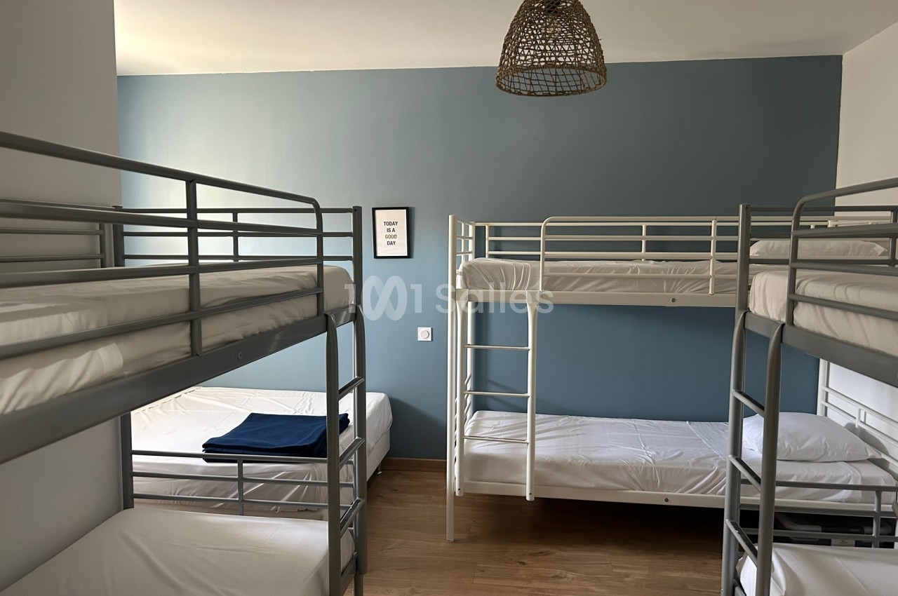 Chambre avec quatre lits superposés en métal, murs bleus et blancs, parquet au sol et suspension en osier.