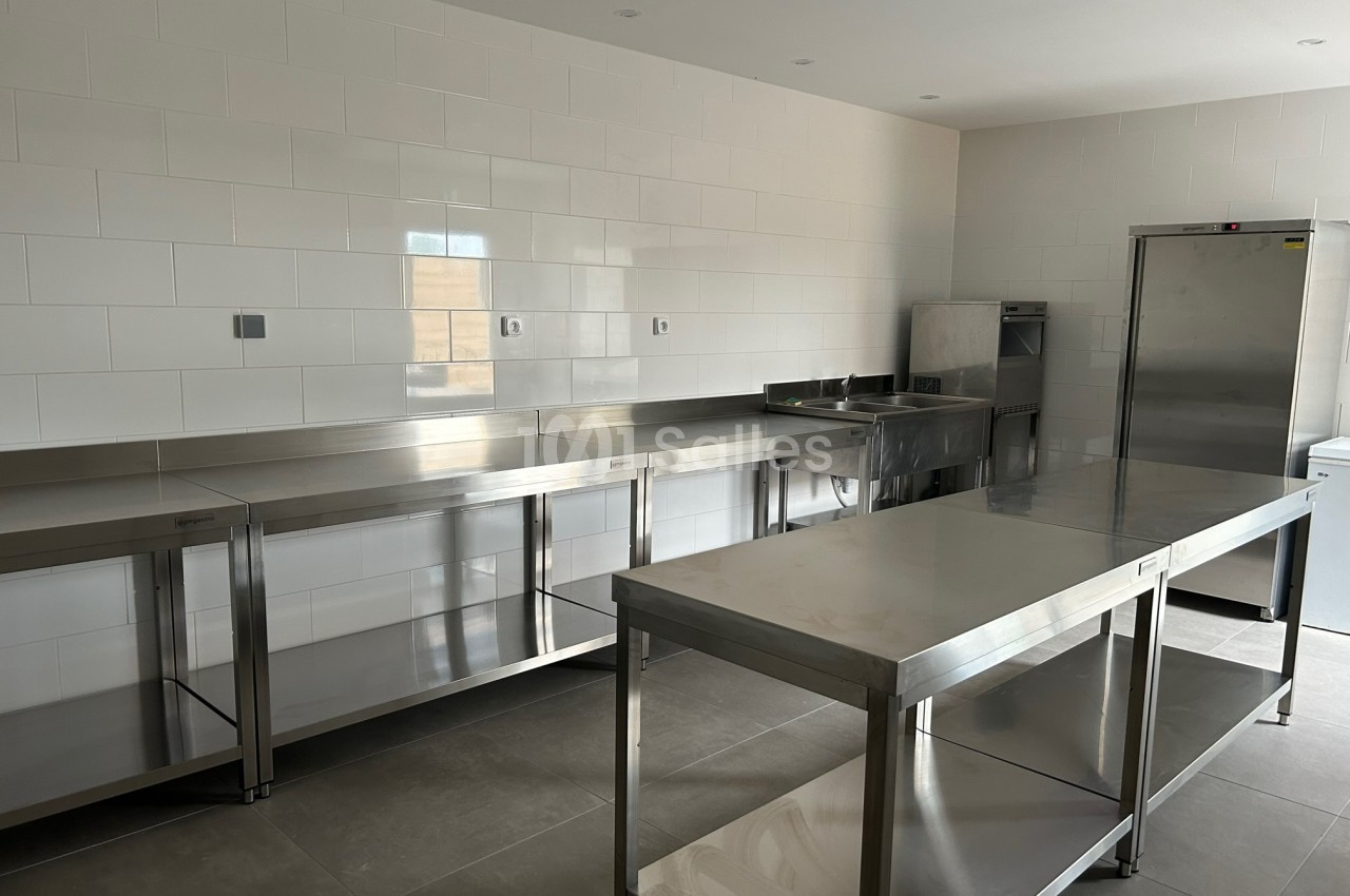 Cuisine professionnelle avec plans de travail en inox, évier, réfrigérateur et murs carrelés blancs.