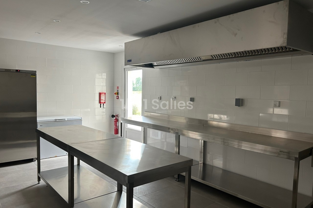 Cuisine professionnelle avec plans de travail en inox, hotte d'extraction, réfrigérateur et murs carrelés blancs.