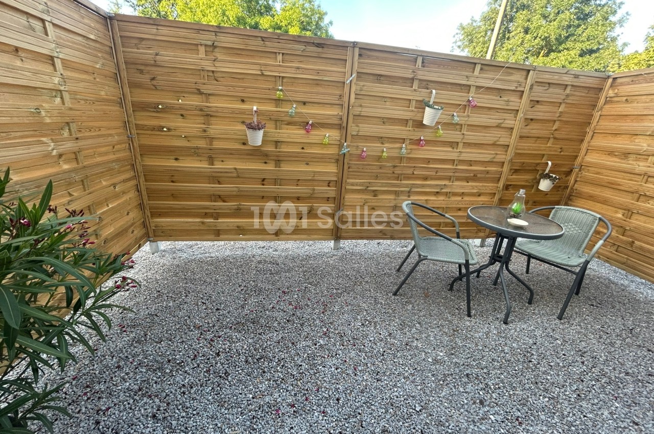 Terrasse en gravier avec table, chaises en métal, clôture en bois décorée de pots suspendus et plantes environnantes.