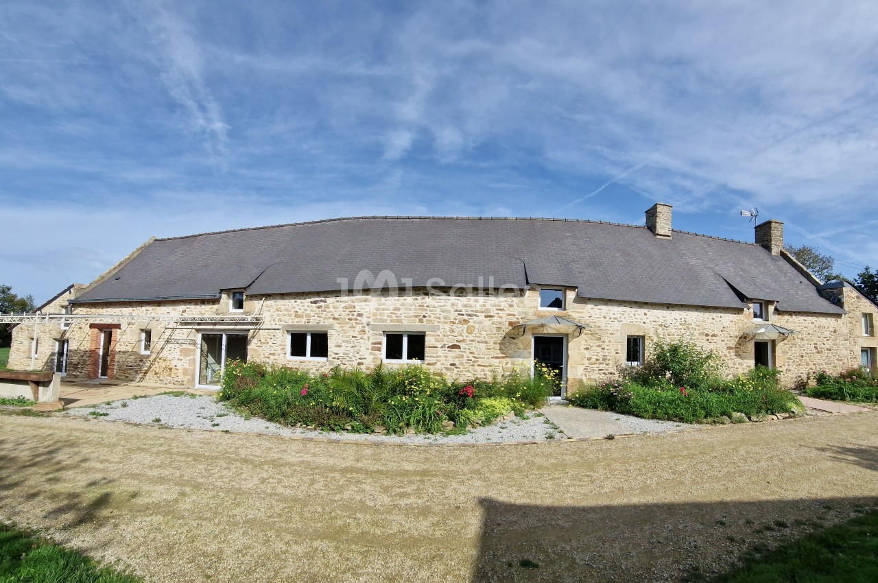 Façade d'une longère en pierre avec toit en ardoise, entourée de jardins fleuris et d'une allée gravillonnée.