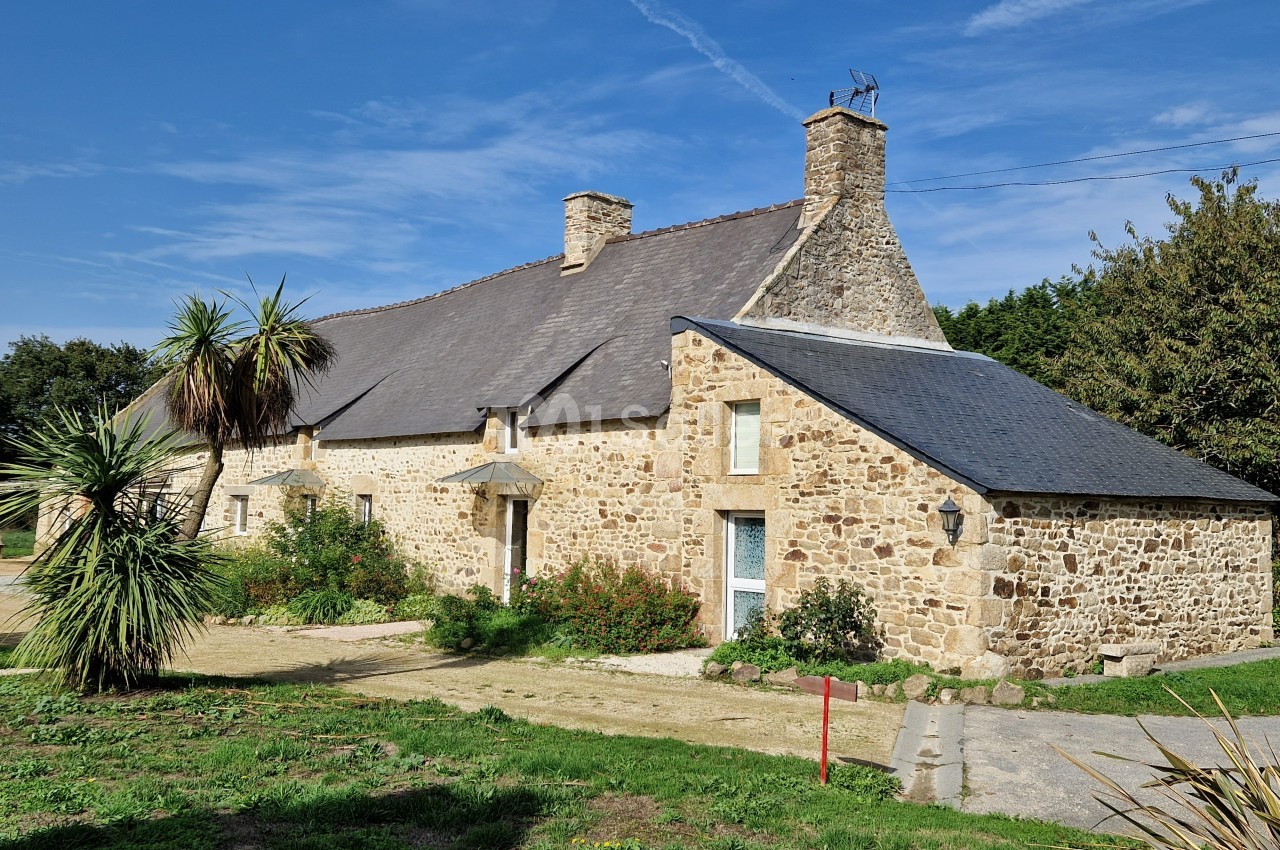Maison en pierre traditionnelle avec toit en ardoise, entourée de végétation et d'un ciel dégagé.