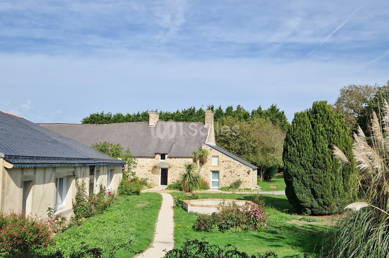 Bâtisse en pierre avec toit en ardoise entourée de verdure, jardin et petit bassin sous un ciel dégagé.