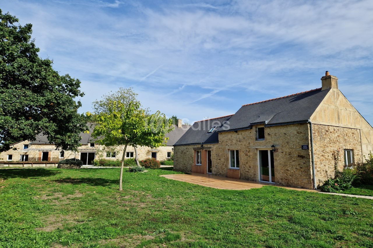 Bâtiments en pierre avec toits en ardoise entourés de pelouse et d'arbres sous un ciel bleu.