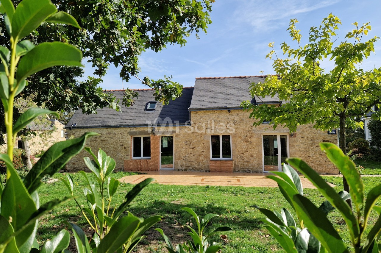 Façade d'une maison en pierre avec toit en ardoise, entourée de verdure et éclairée par un ciel ensoleillé.
