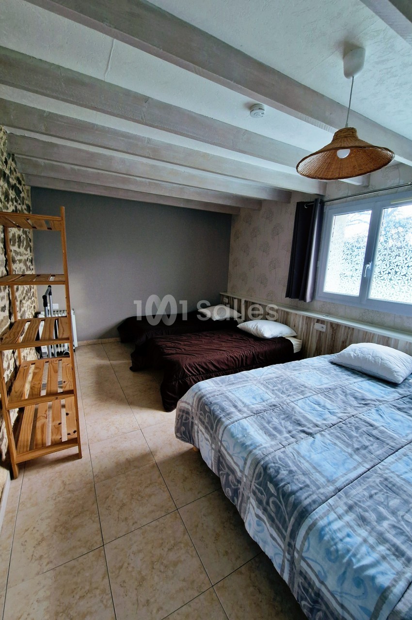 Chambre avec deux lits doubles, étagère en bois, mur en pierre et grande fenêtre laissant entrer la lumière naturelle.