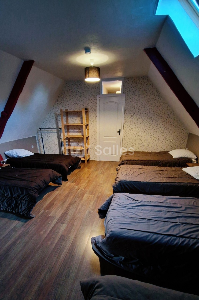 Dortoir mansardé avec six lits simples alignés, parquet au sol, étagère en bois et lumière naturelle d'une fenêtre de toit.