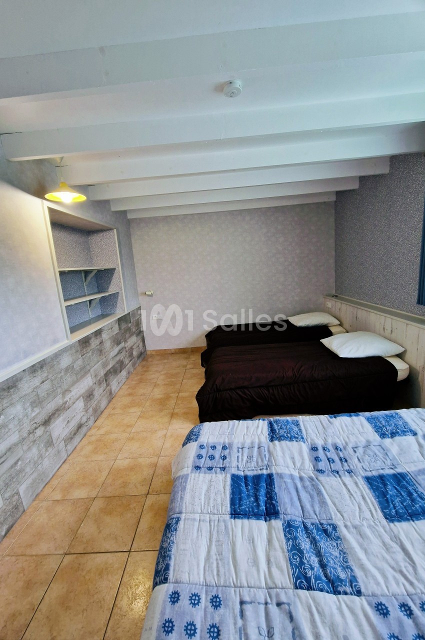 Chambre avec trois lits simples, sol en carrelage, murs clairs, étagères intégrées et plafond avec poutres apparentes.