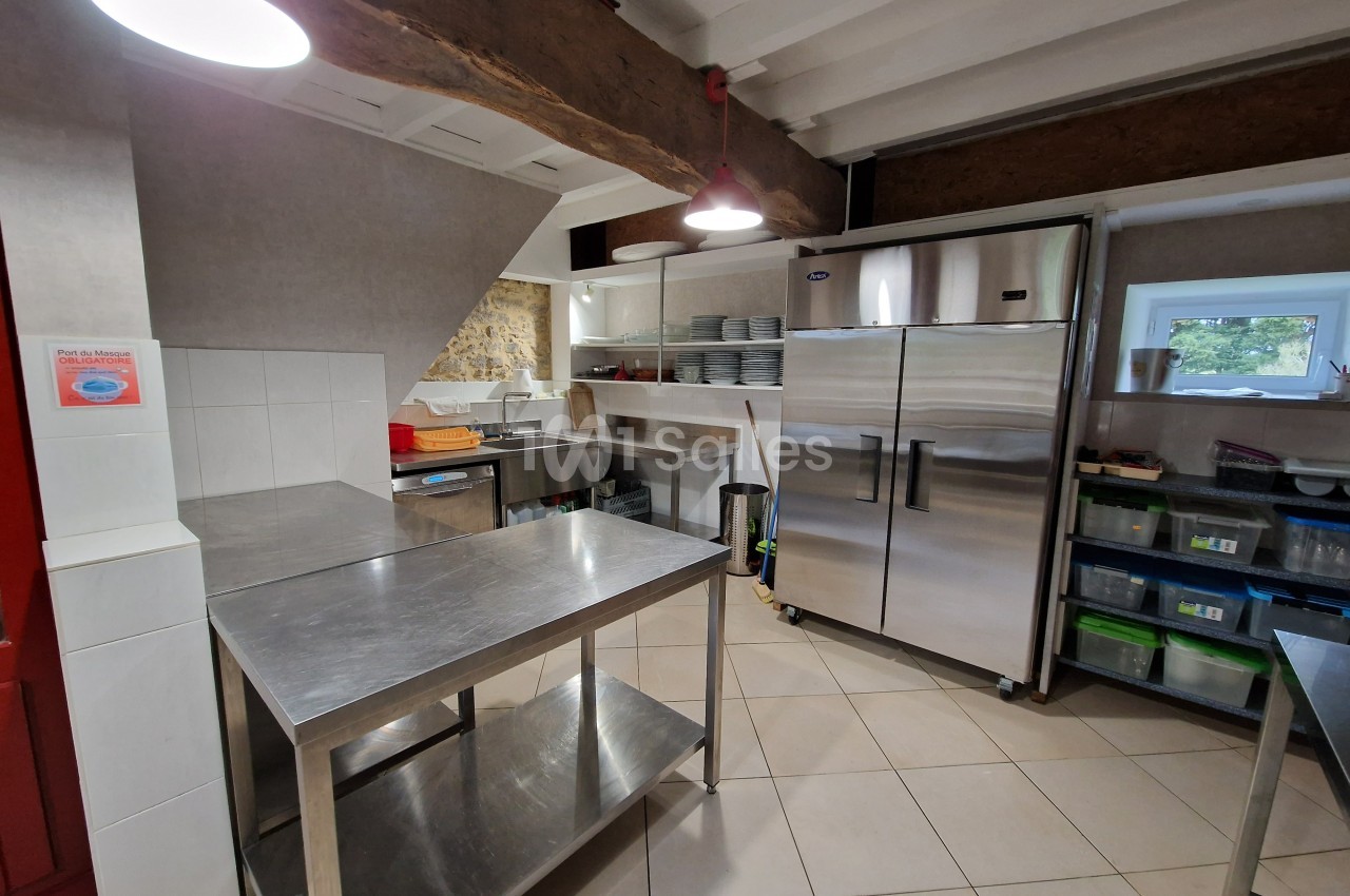 Cuisine professionnelle avec plans de travail en inox, réfrigérateur, étagères de rangement et éclairage suspendu.