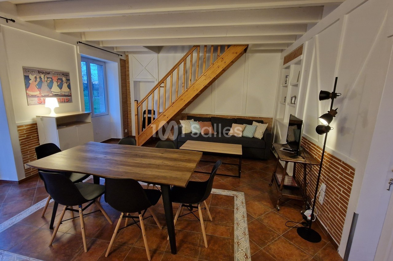 Salle à manger avec table en bois, chaises noires, canapé, escalier en bois et éclairage chaleureux.