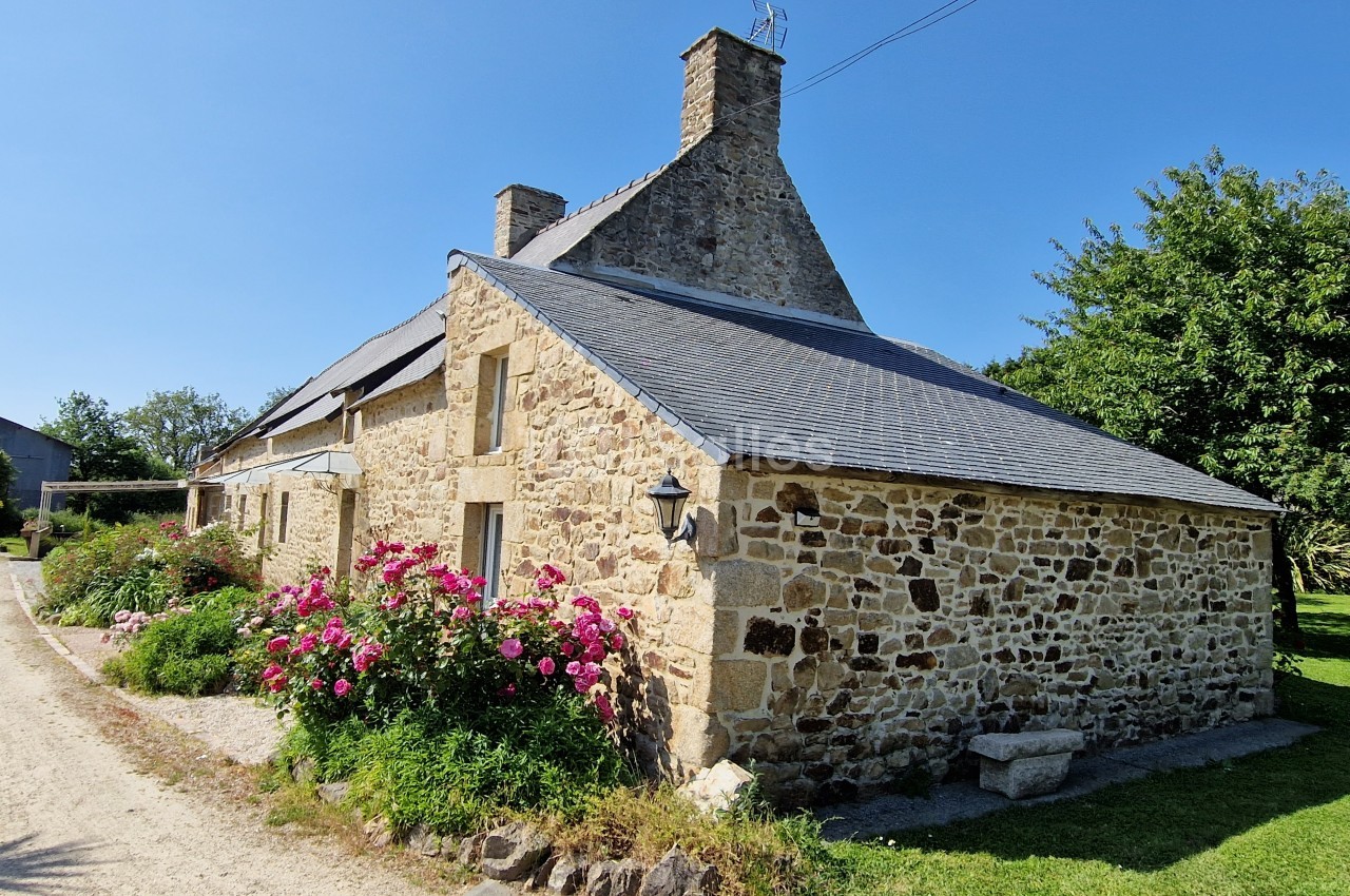 Maison en pierre traditionnelle avec toit en ardoise, entourée de fleurs et d'arbres sous un ciel dégagé.