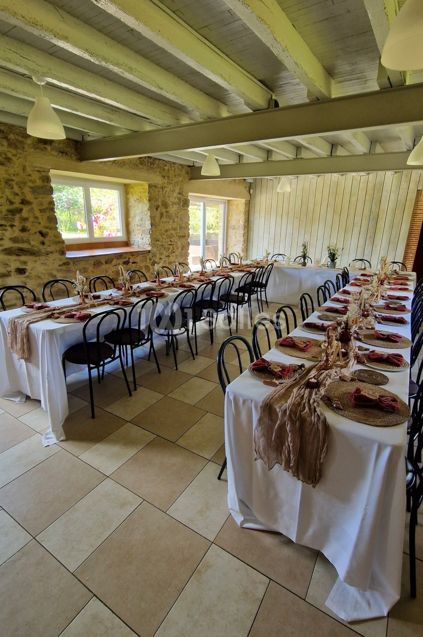 Salle de réception avec tables alignées, nappes blanches, décorations rustiques et lumière naturelle provenant des fenêtres.