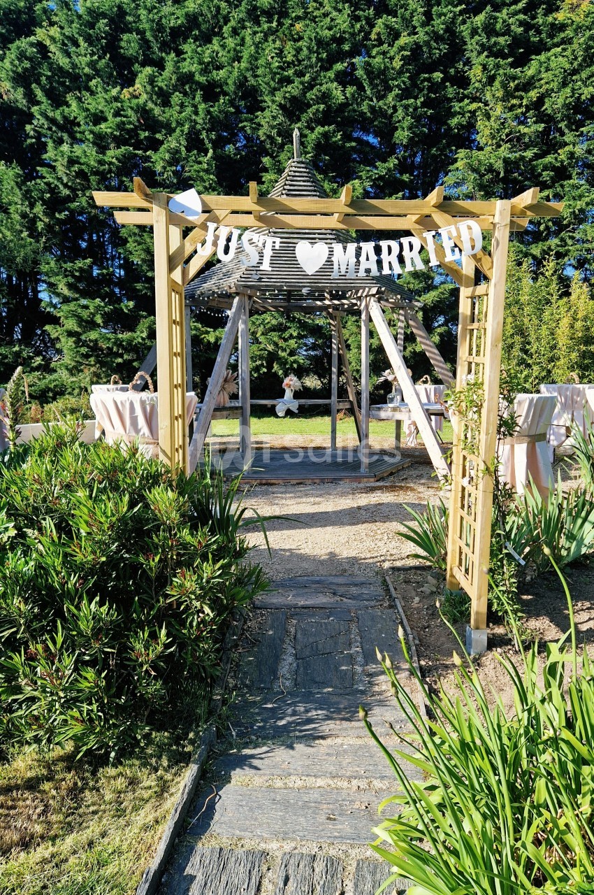 Entrée d'une pergola en bois décorée d'une bannière ’Just Married’, entourée de verdure et de tables dressées.