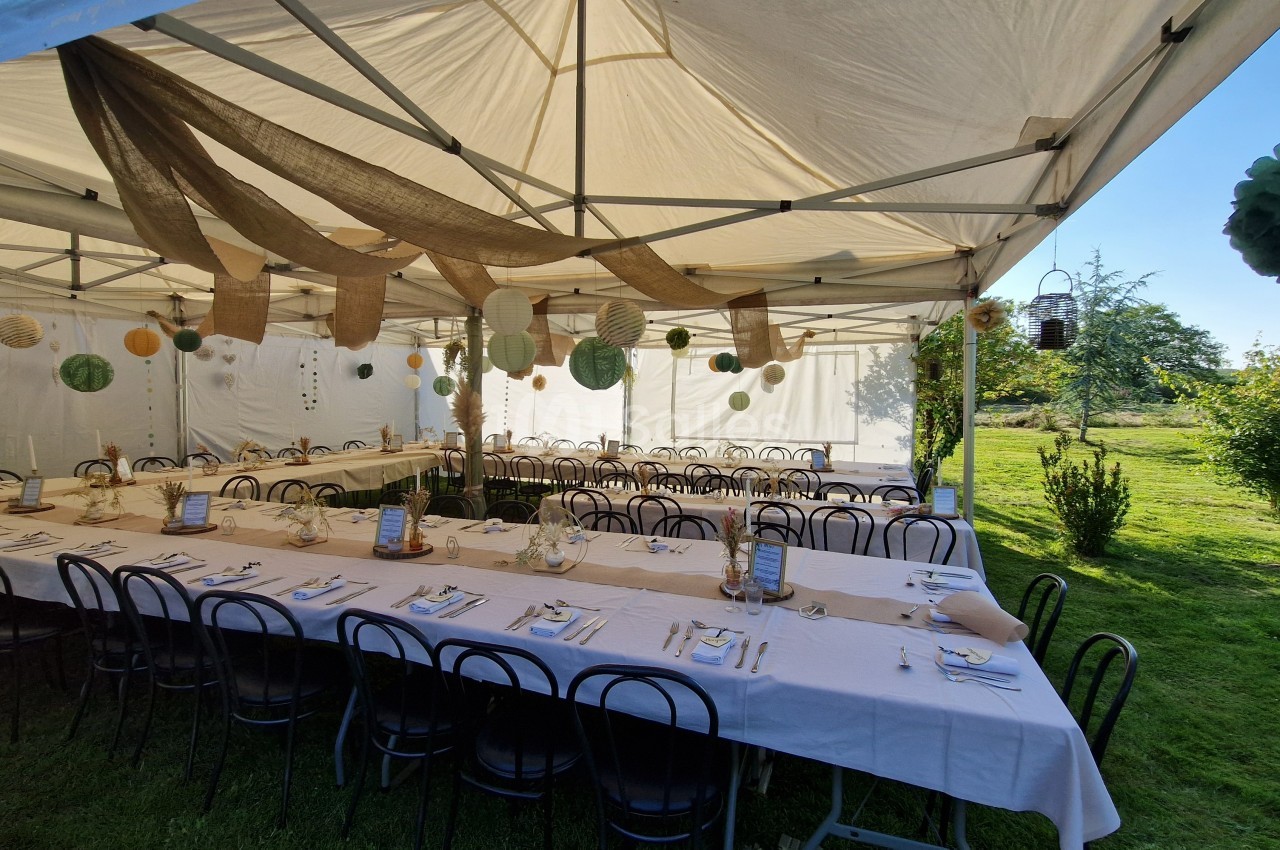 Tables dressées sous une tente blanche dans un jardin, décorées de lanternes suspendues et de nappes blanches.