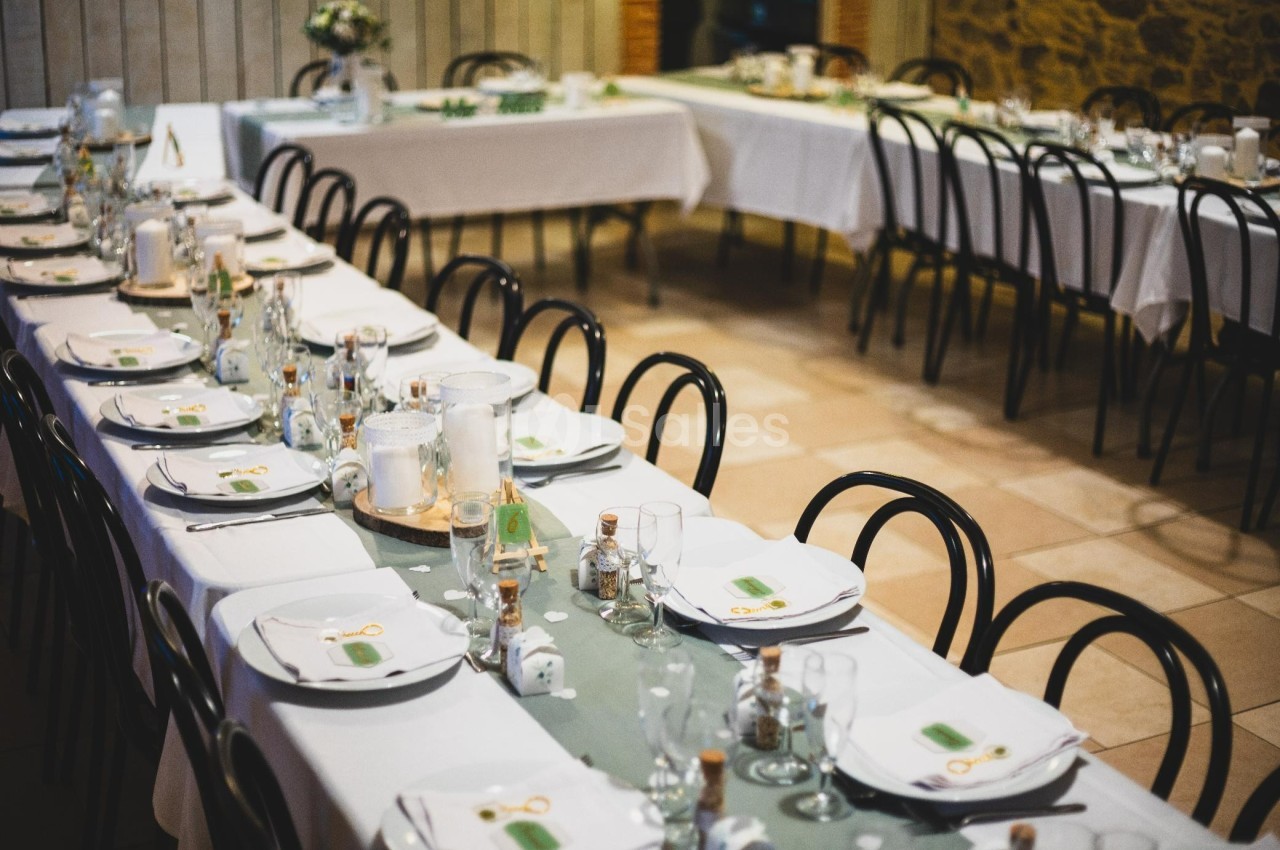 Tables dressées avec nappes blanches, vaisselle et décorations, disposées dans une salle de réception lumineuse.