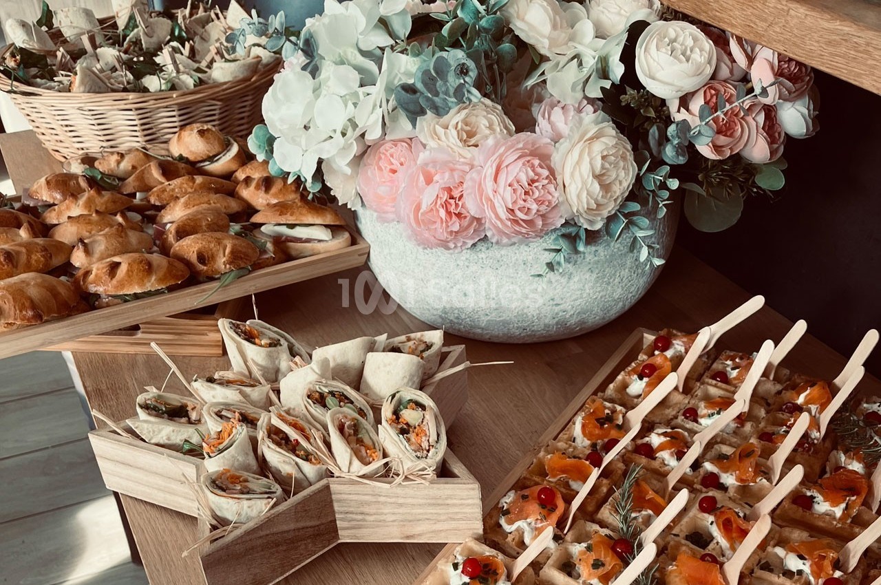 Plateau de bouchées apéritives variées avec wraps, feuilletés et canapés, décoré de fleurs en arrière-plan.