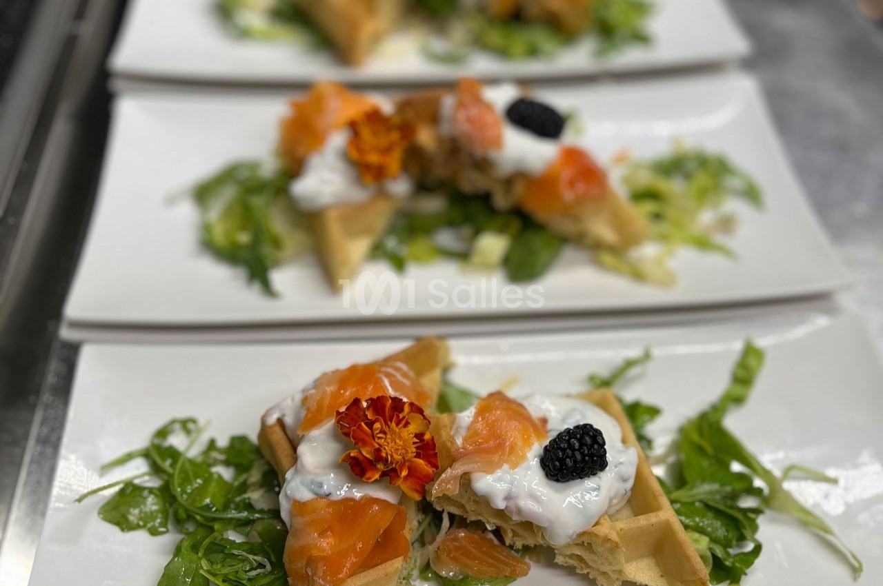 Assiette de gaufres garnies de saumon fumé, crème, mûres et fleurs comestibles, servies sur un lit de salade verte.