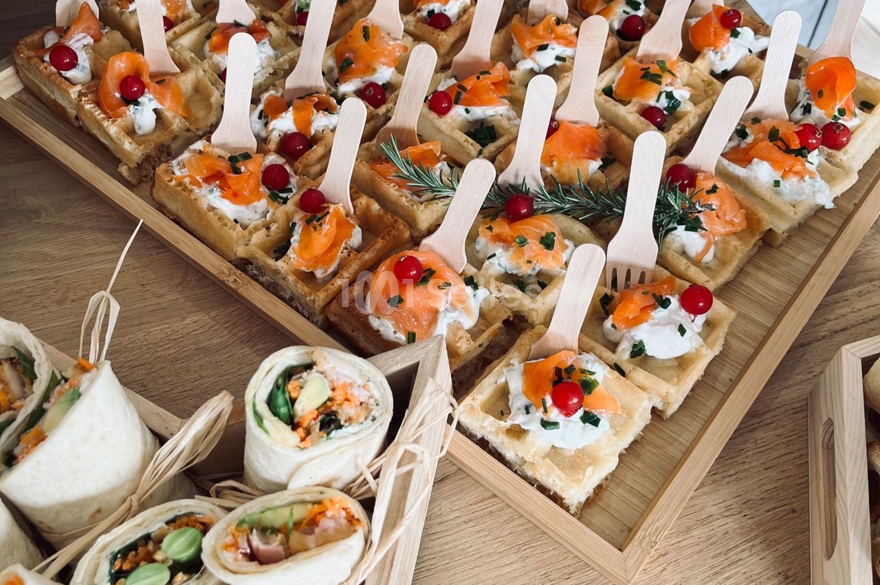 Plateau de bouchées apéritives comprenant wraps garnis et gaufres salées au saumon et fromage frais, décorées de baies…
