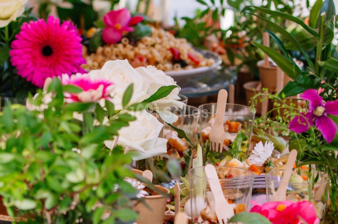 Table garnie de fleurs colorées, de salades en verrines et d'autres plats entourés de verdure.