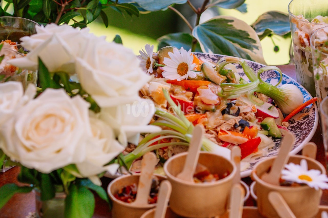 Assiette garnie de légumes colorés et petits plats en bois, entourés de fleurs blanches et de verdure.