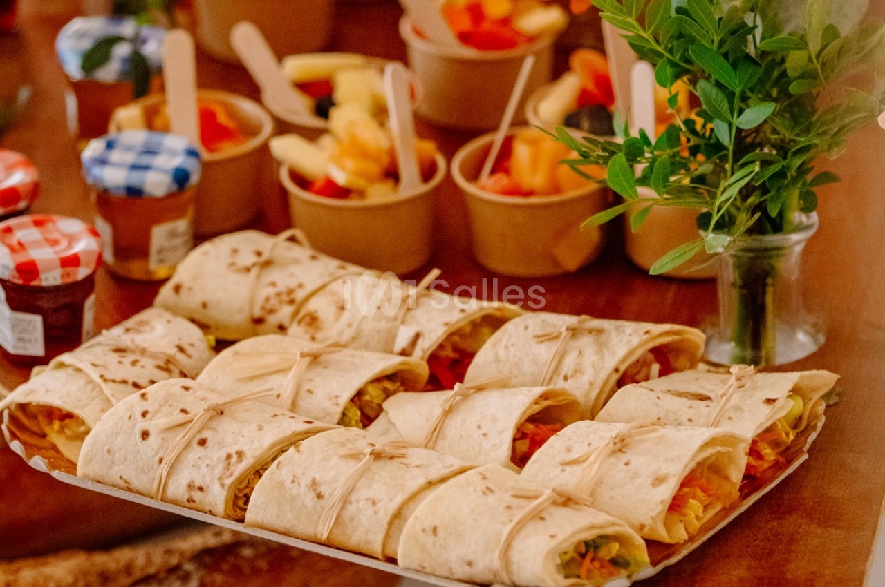 Plateau de wraps garnis de légumes, entouré de pots de fruits et d'une plante décorative sur une table en bois.