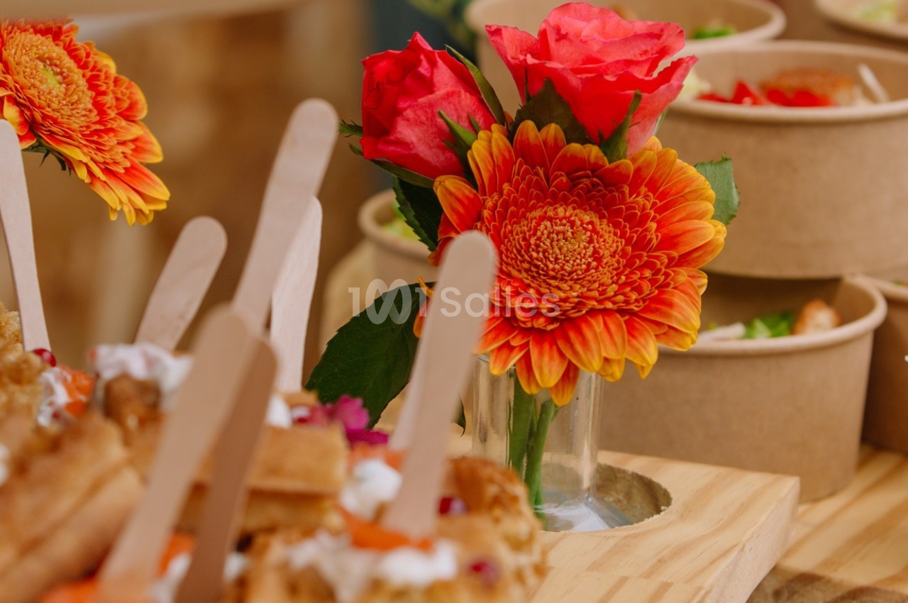 Bouquet de fleurs orange et rouges dans un vase en verre, entouré de plats en carton et de bâtonnets en bois.