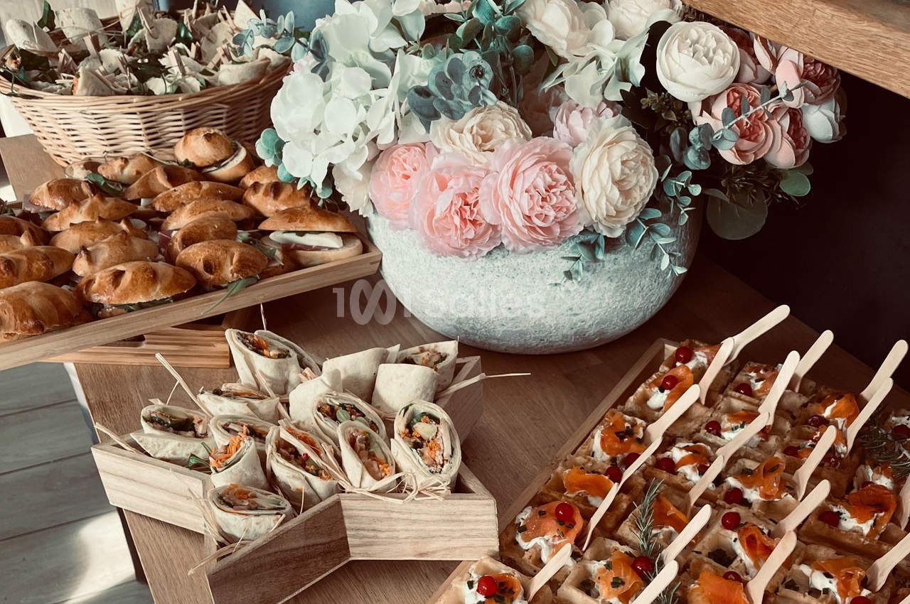 Plateau de bouchées apéritives variées avec wraps, feuilletés et canapés, décoré de fleurs en arrière-plan.