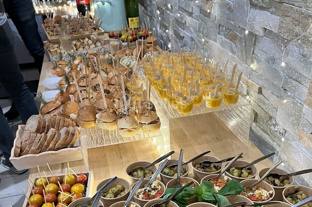 Buffet composé de mini-burgers, verrines, brochettes de tomates, pains et salades, présenté sur des tables en bois.