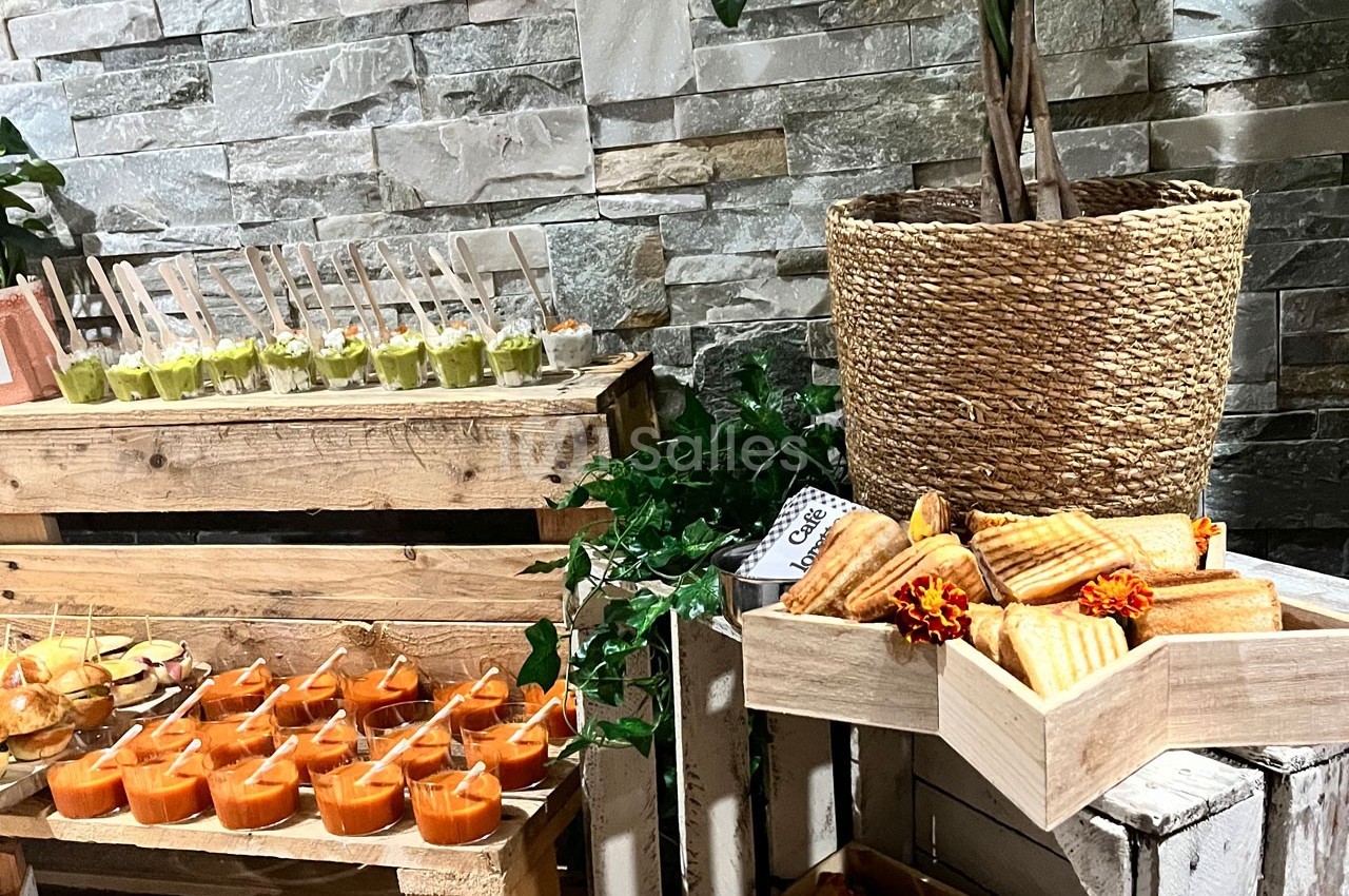 Buffet avec verrines colorées, mini-sandwiches et paninis disposés sur des caisses en bois devant un mur en pierre.