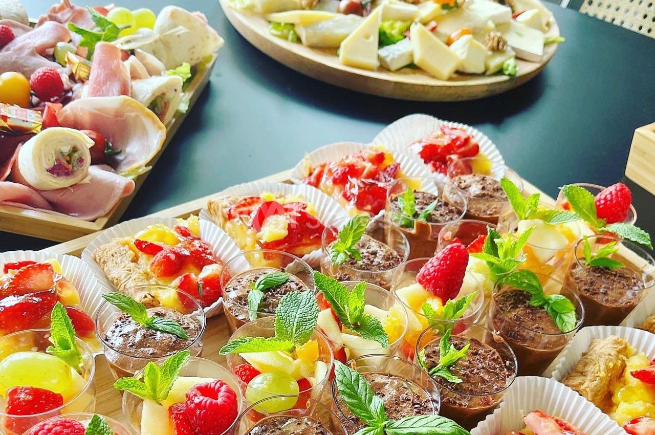 Plateaux de desserts variés avec fruits frais, crèmes et pâtisseries, accompagnés de charcuteries et fromages en arrière…