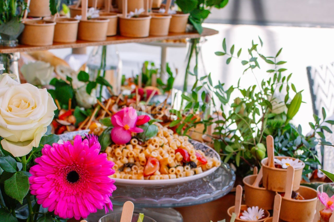 Buffet coloré avec un plat de pâtes décoré de fleurs comestibles, entouré de pots en carton et de verdure.