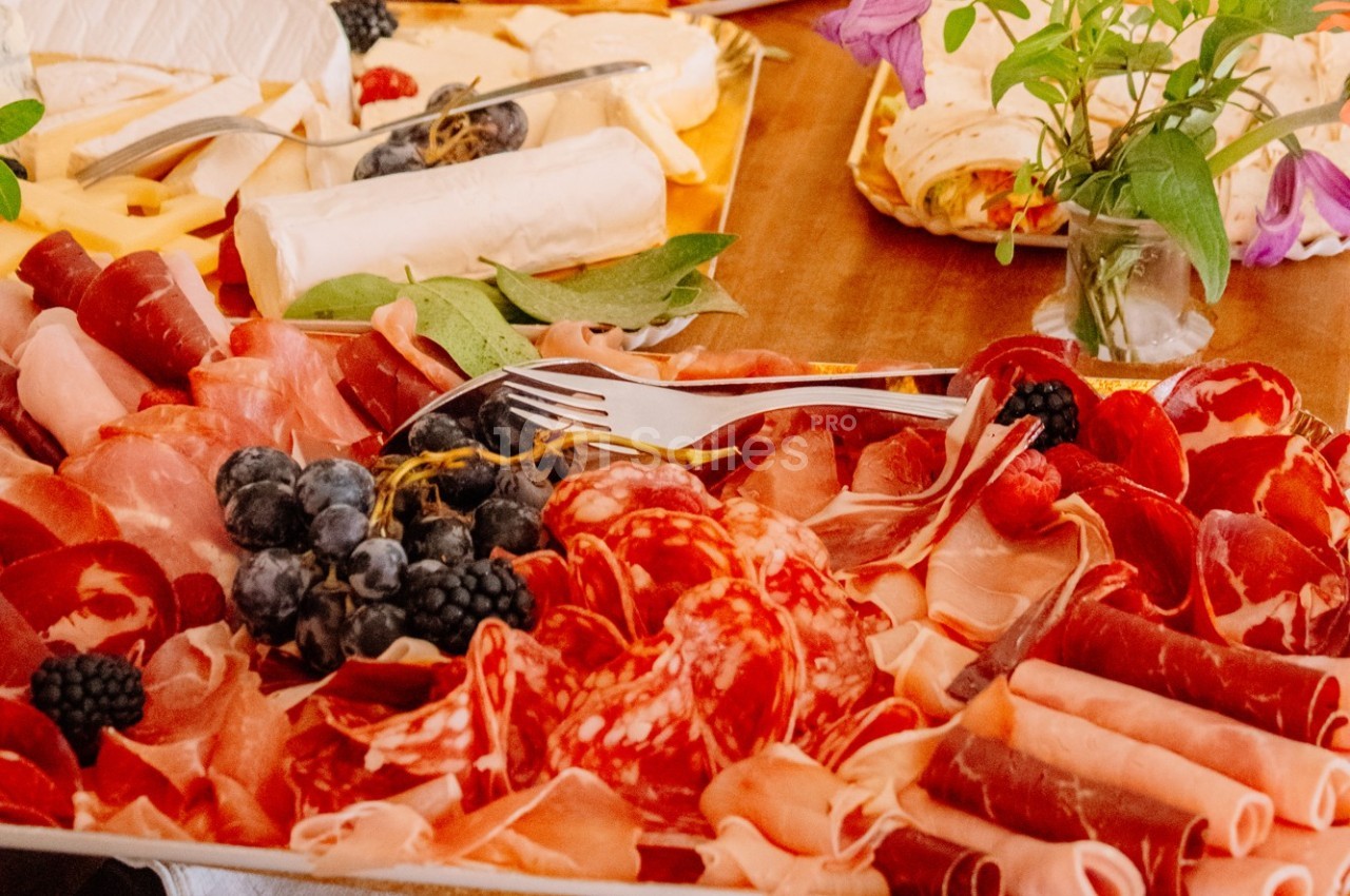 Plateau de charcuterie variée avec jambon, saucisson, fruits rouges et fromage, décoré de feuilles et fleurs.