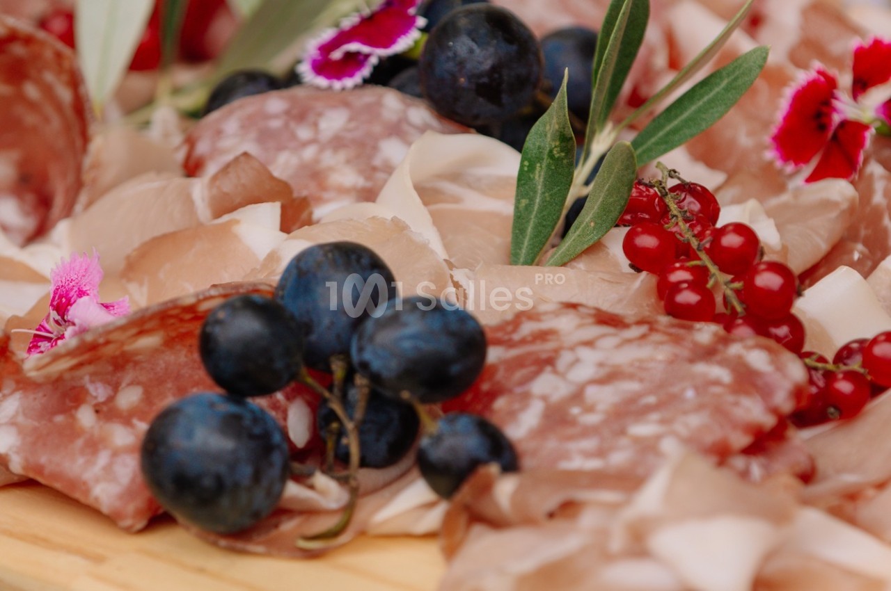 Plateau de charcuterie avec tranches de saucisson, jambon, raisins noirs, groseilles rouges et décorations florales.