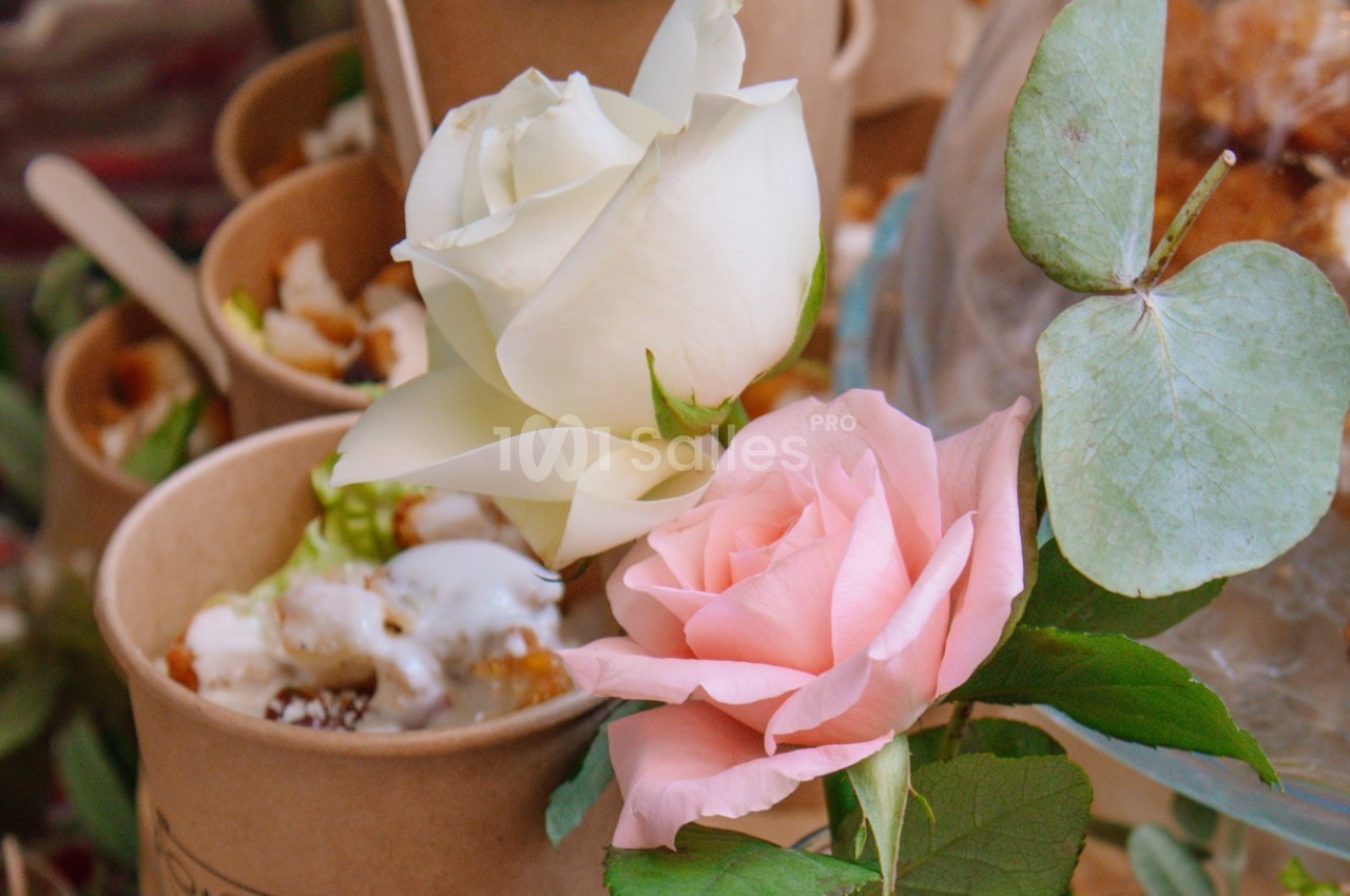 Deux roses, blanche et rose, posées près de feuilles vertes, devant des pots en carton contenant des desserts.