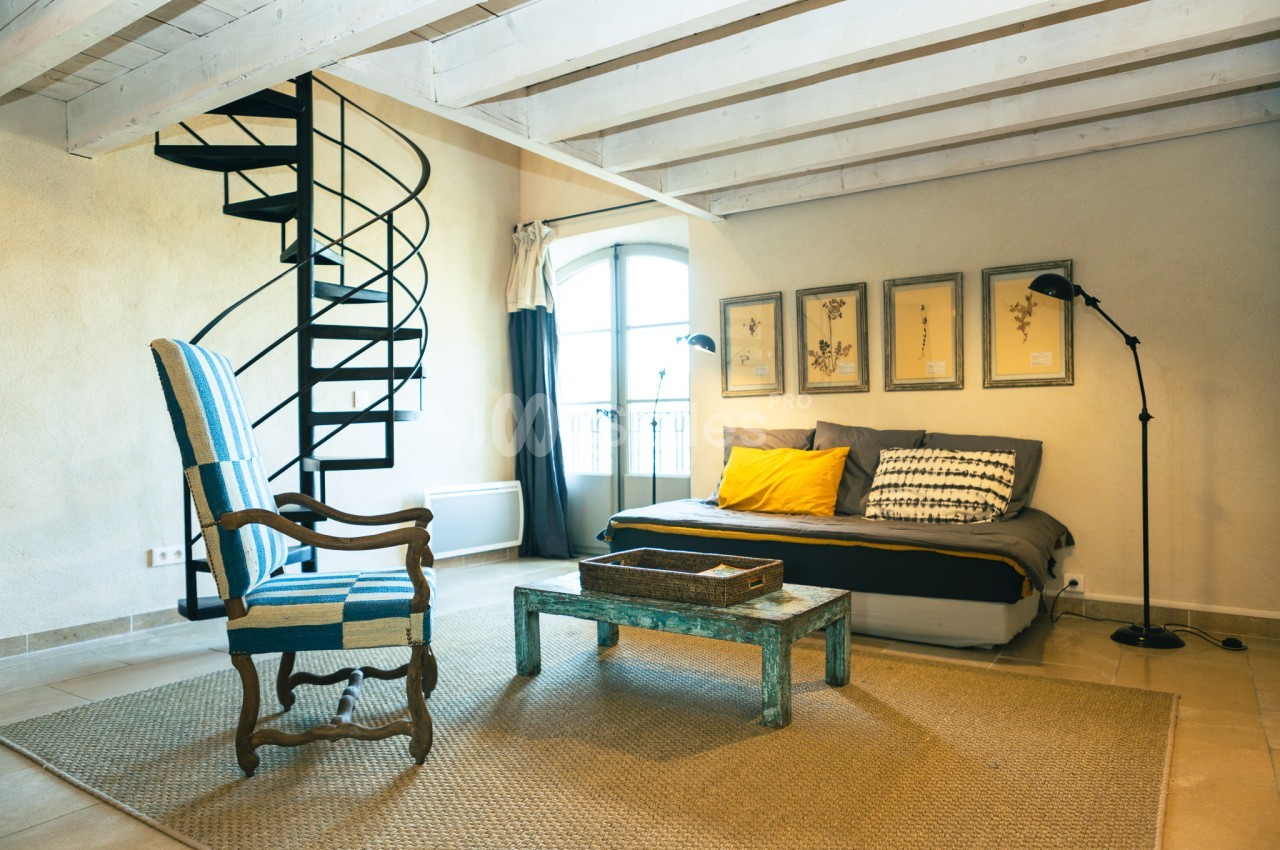 Salon lumineux avec fauteuil, canapé, table basse en bois, escalier en colimaçon et décoration murale.