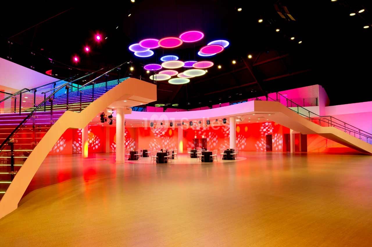 Salle spacieuse avec éclairage coloré au plafond, escaliers modernes et tables dispersées sur un sol clair.