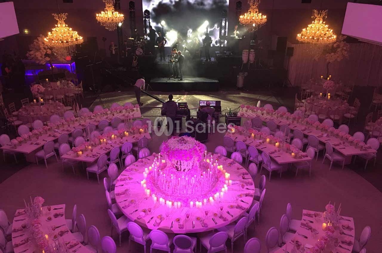 Salle de réception décorée avec des tables rondes, des chandeliers lumineux et une scène avec un écran en arrière-plan.