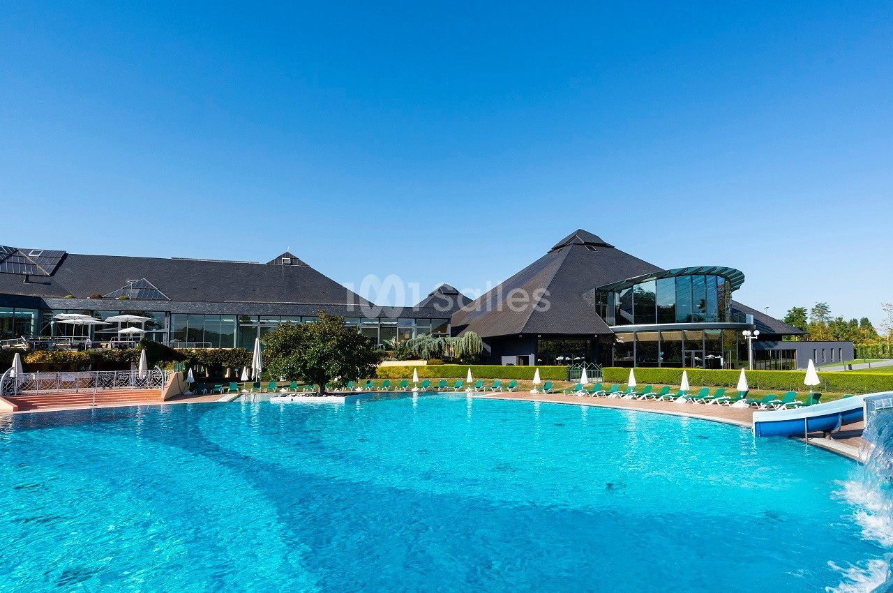Piscine extérieure avec eau turquoise entourée de chaises longues, devant un bâtiment moderne sous un ciel bleu.