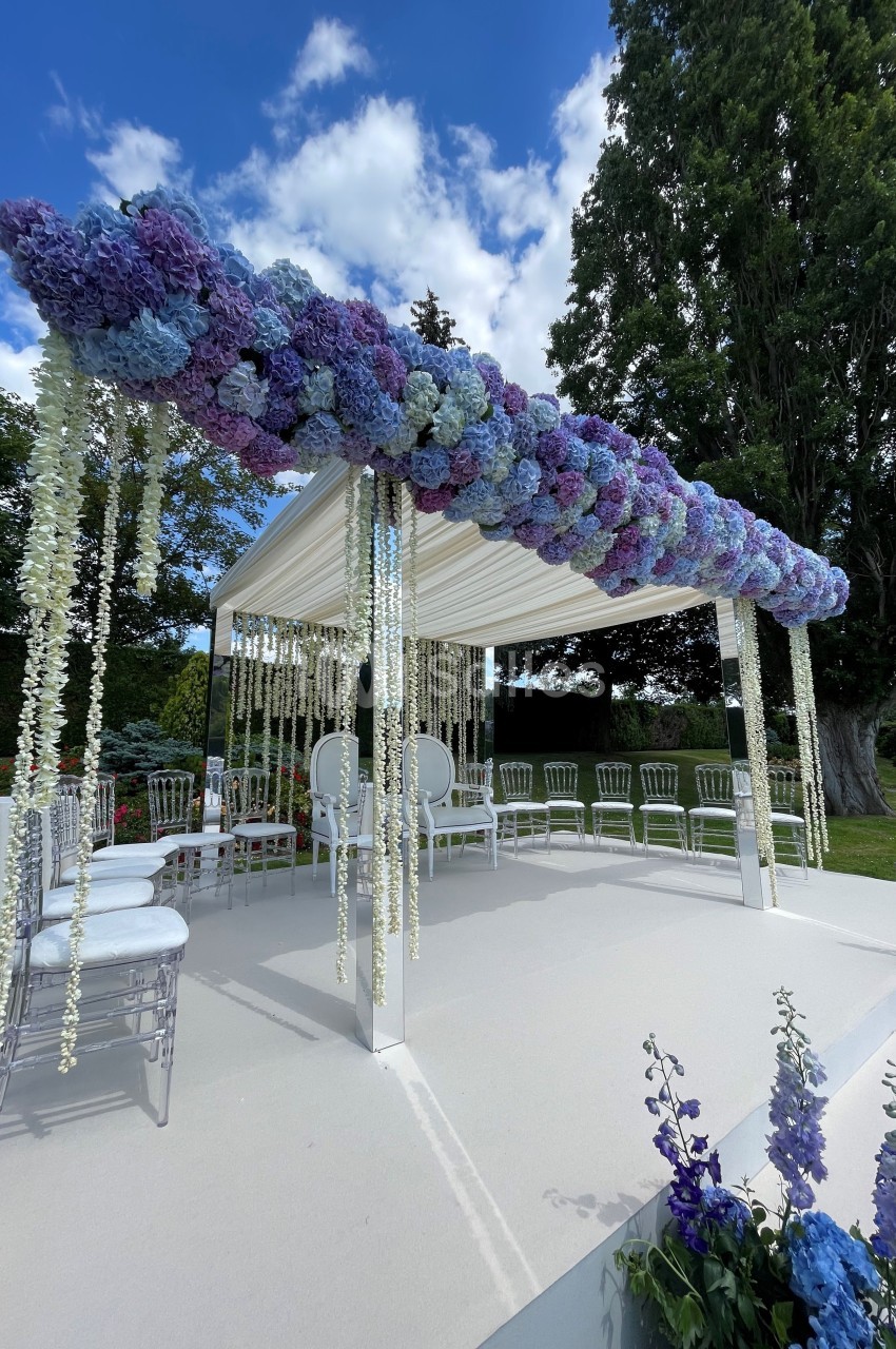 Structure de cérémonie décorée de fleurs violettes et blanches, entourée de chaises, dans un jardin sous un ciel bleu.
