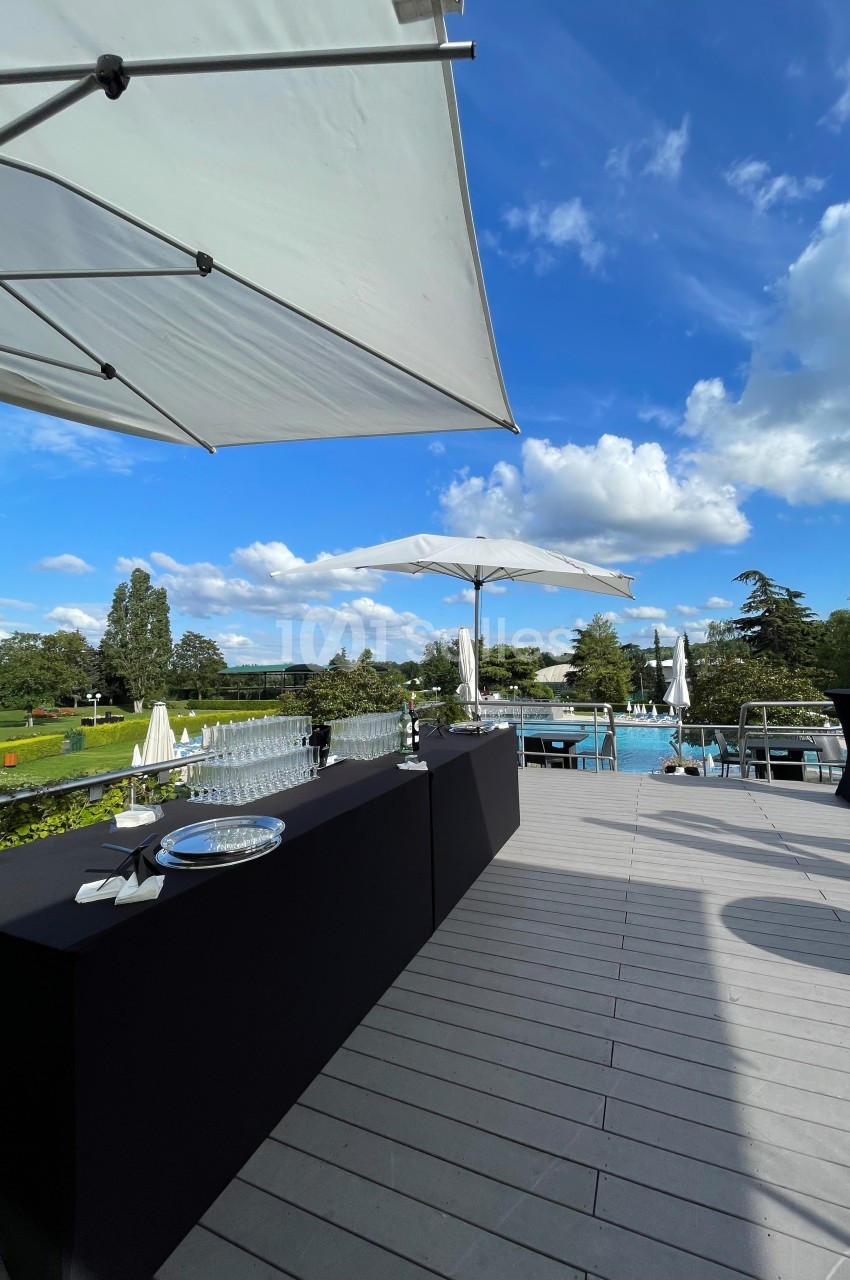 Terrasse ensoleillée avec tables noires dressées, parasols blancs et vue sur une piscine entourée de verdure.