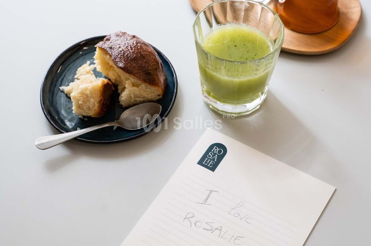 Un verre de jus vert, une brioche sur une assiette avec une cuillère, et une note manuscrite sur une table blanche.