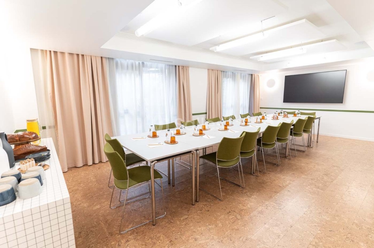 Salle de réunion lumineuse avec une grande table rectangulaire, chaises vertes, écran mural et rideaux beiges.