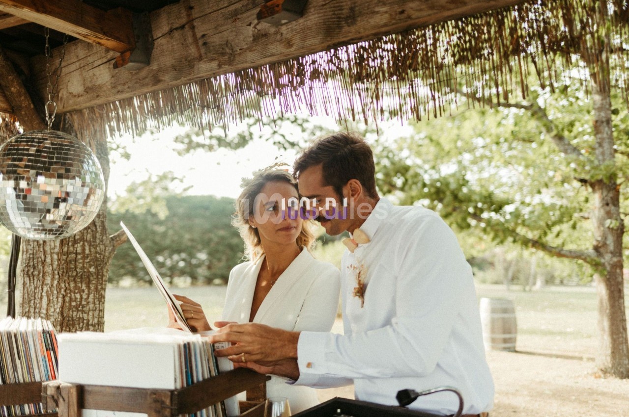 Un couple examine des disques vinyles sous un abri en bois, entouré de verdure et d'une boule à facettes suspendue.